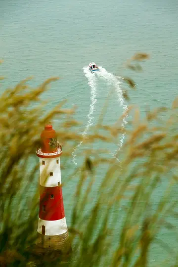 Un faro rojo y blanco se alza en el mar turquesa, parcialmente oculto por juncos dorados y verdes desenfocados en primer