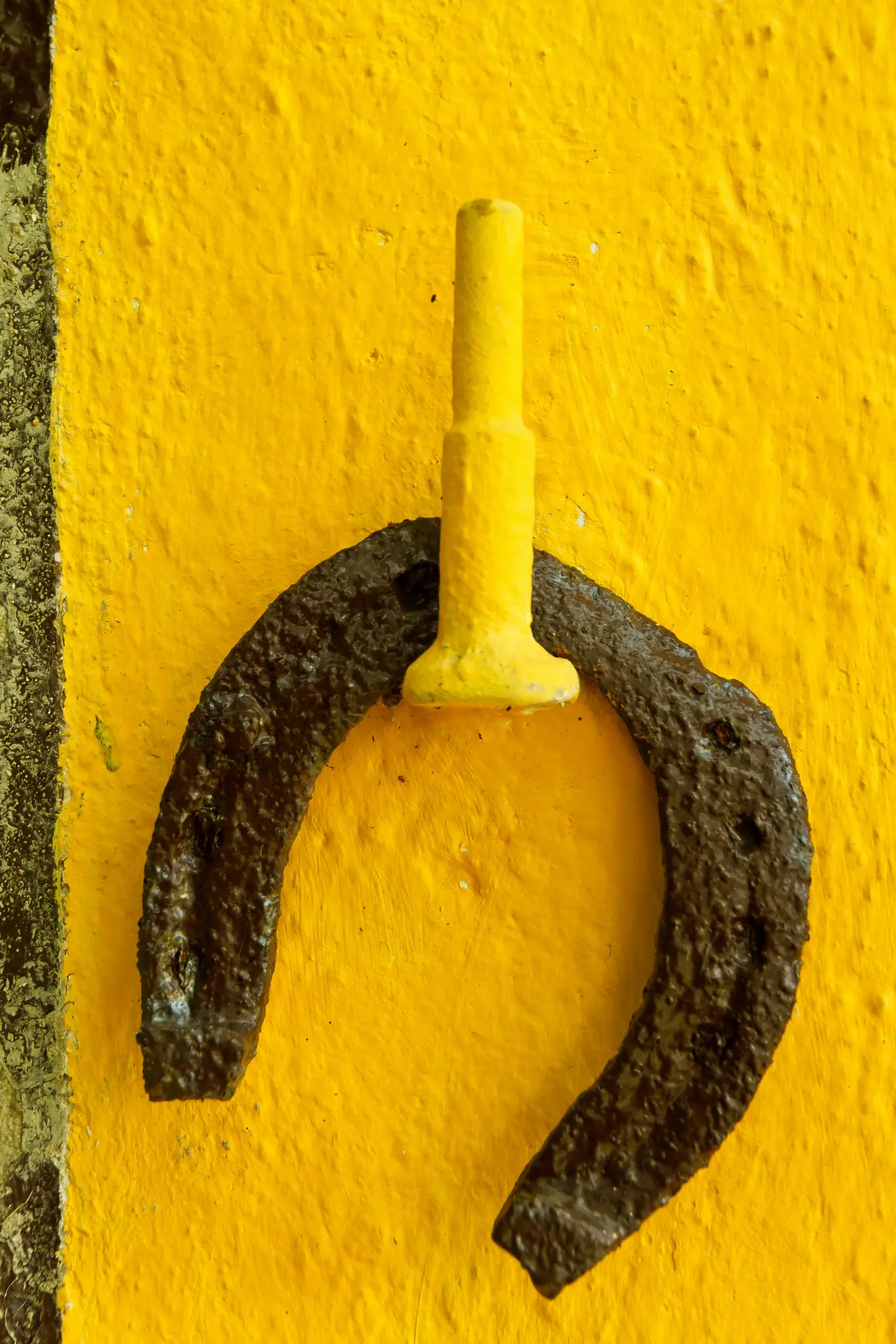 Ein dunkles, rostiges Hufeisen hängt kopfüber an einer hellen, gelben, strukturierten Wand und ist an einem kurzen gelben