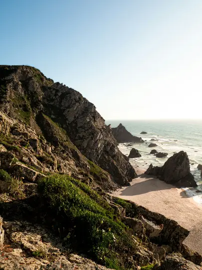 Vista dall'alto di una costa aspra con ripide scogliere rocciose coperte da vegetazione verde, una spiaggia sabbiosa e…