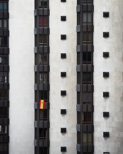 Una bandiera spagnola, rossa e gialla, pende da una finestra con cornice scura su un edificio alto con una facciata beige