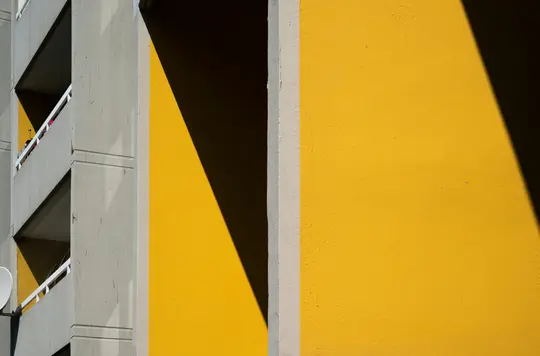Una pared amarilla vibrante con una sombra triangular oscura y afilada a la derecha, junto a la fachada de un edificio de