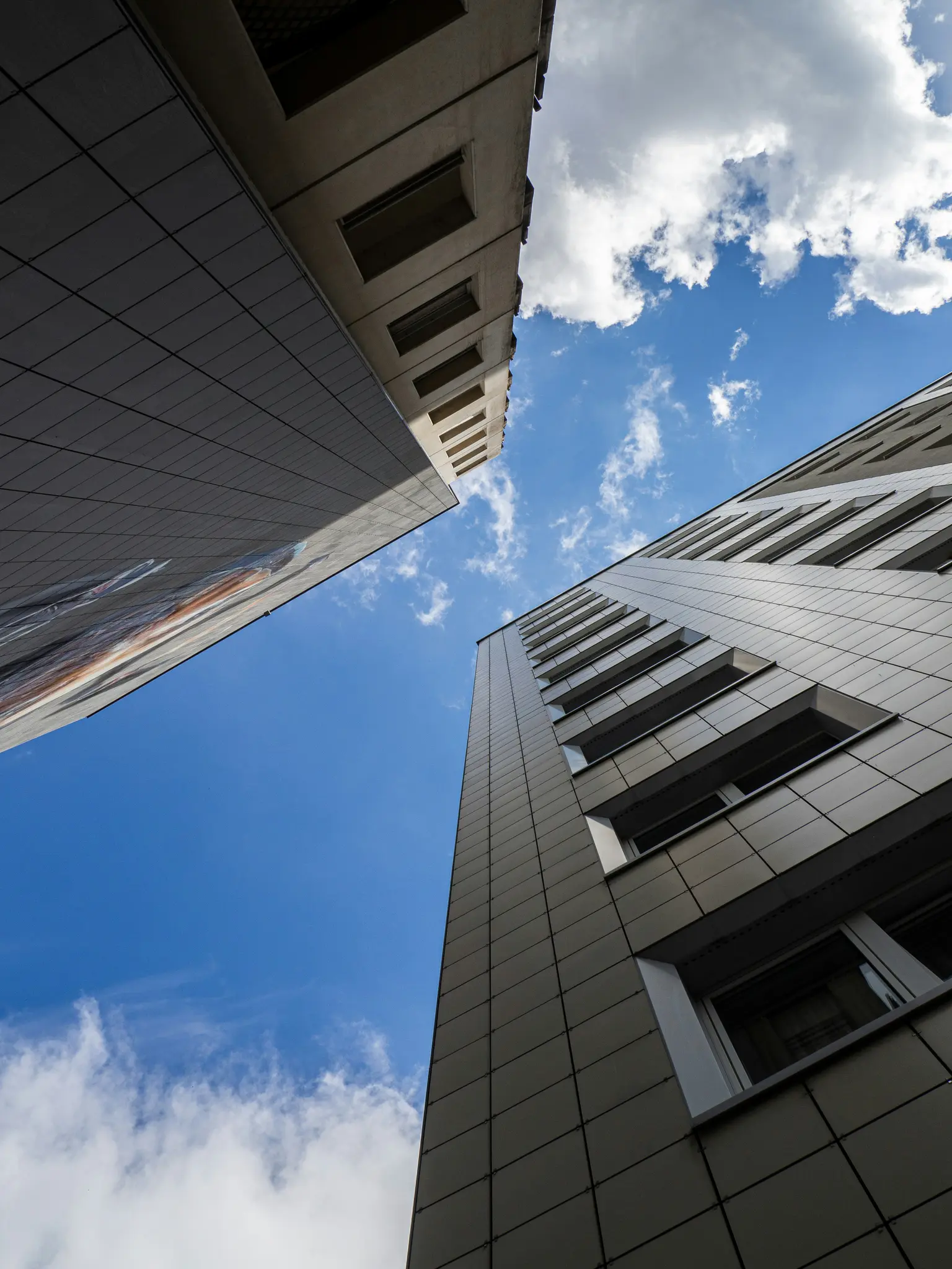 A low-angle shot of three tall buildings with modern facades against a bright blue sky with scattered white clouds.