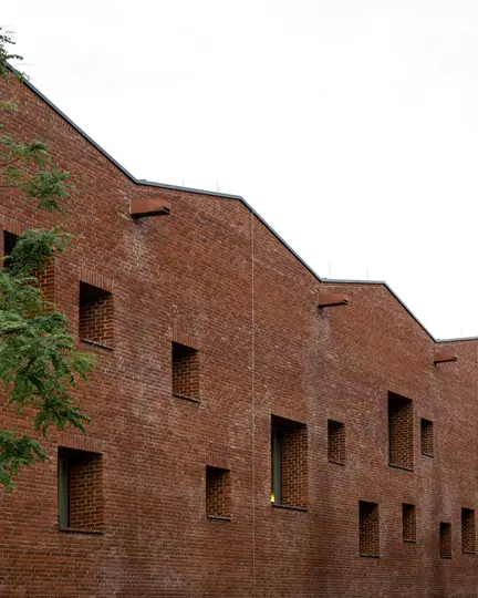 Ein modernes Gebäude mit einer rotbraunen Backsteinfassade und einer gestuften Dachlinie vor einem weißen Himmel.
