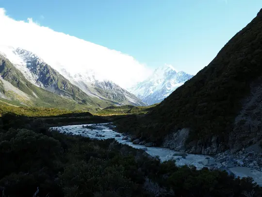 Una toma amplia de un río glacial de color azul claro que serpentea por un valle. Montañas nevadas se elevan a la izquierda