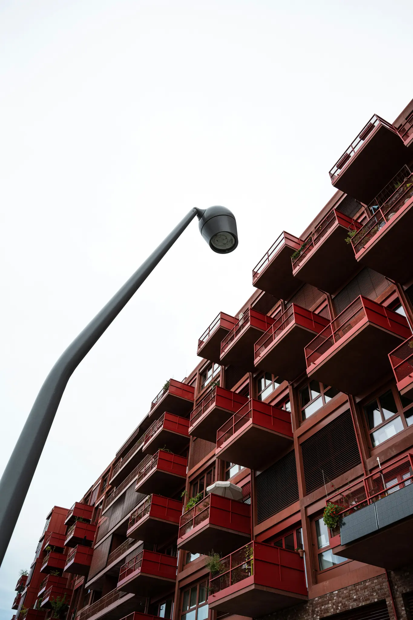 Pohľad z nízkeho uhla na modernú budovu s početnými červenými balkónmi a šedým pouličným svetlom zakrivujúcim sa do záberu.