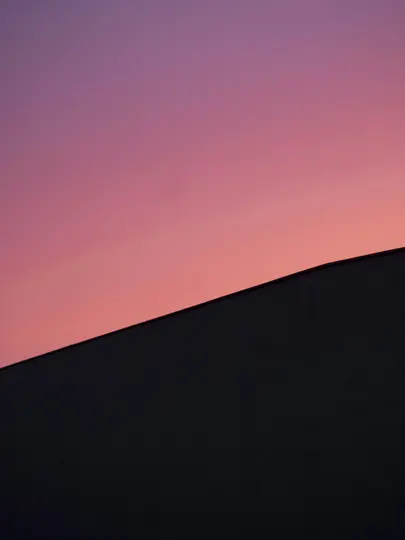 Una foto minimalista che mostra la silhouette scura e angolare di un edificio contro un cielo che sfuma dal viola intenso a…