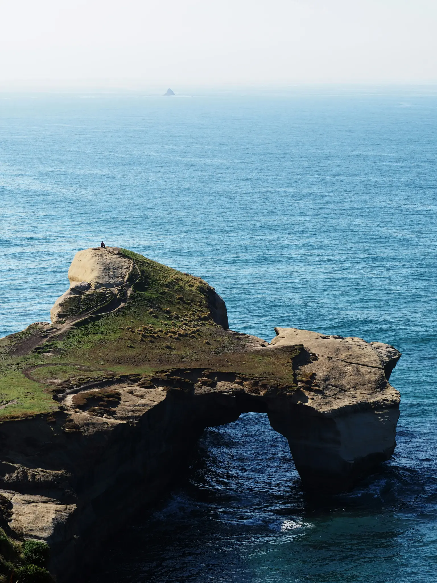 Una persona se sienta en lo alto de un acantilado cubierto de hierba con vistas a un arco de roca natural. El arco se