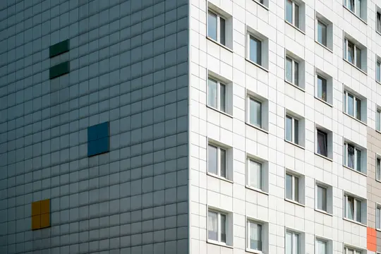 Una moderna facciata di edificio divisa da luce e ombra. Il lato sinistro è grigio scuro con quadrati verdi, blu e arancioni.