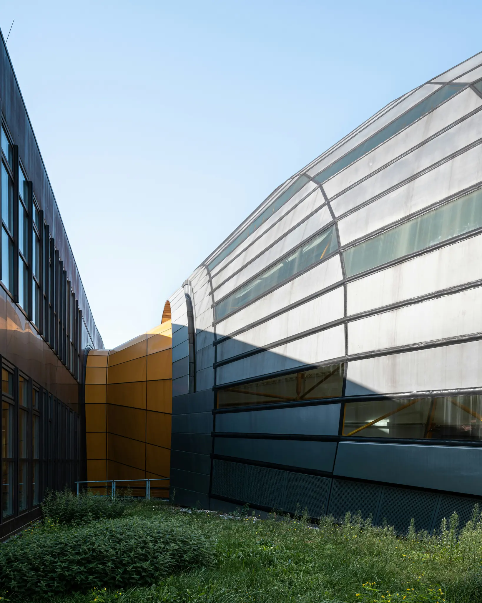 Moderný architektonický komplex s tmavou budovou s obdĺžnikovými oknami, oranžovou zakrivenou konštrukciou a sivou…