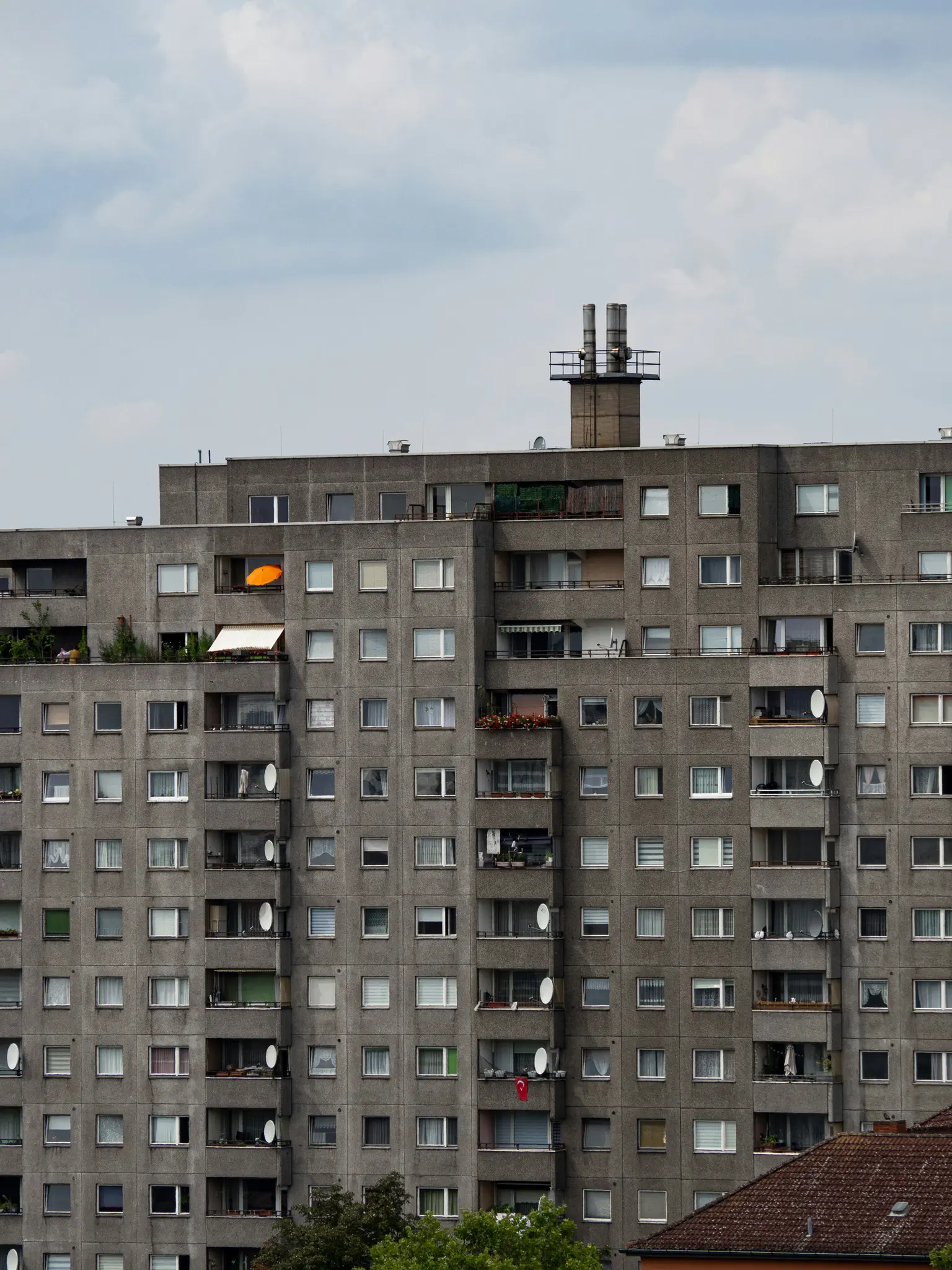 Vysoký, sivý betónový bytový dom vypĺňa rám, s mnohými oknami a balkónmi pod zamračenou oblohou.
