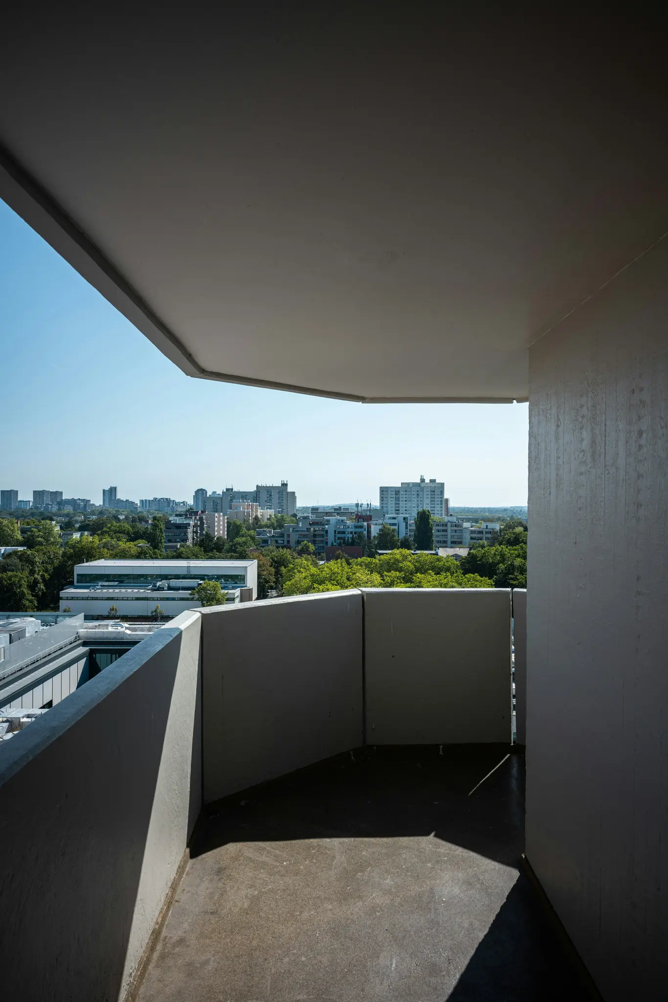 Un balcón de hormigón con vistas a una ciudad. El primer plano muestra el suelo y la barandilla del balcón, con sombras. Al