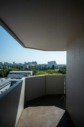 Ein Betonbalkon mit Blick auf eine Stadt. Der Vordergrund zeigt den Balkonboden und die Geländer, mit Schatten. Im