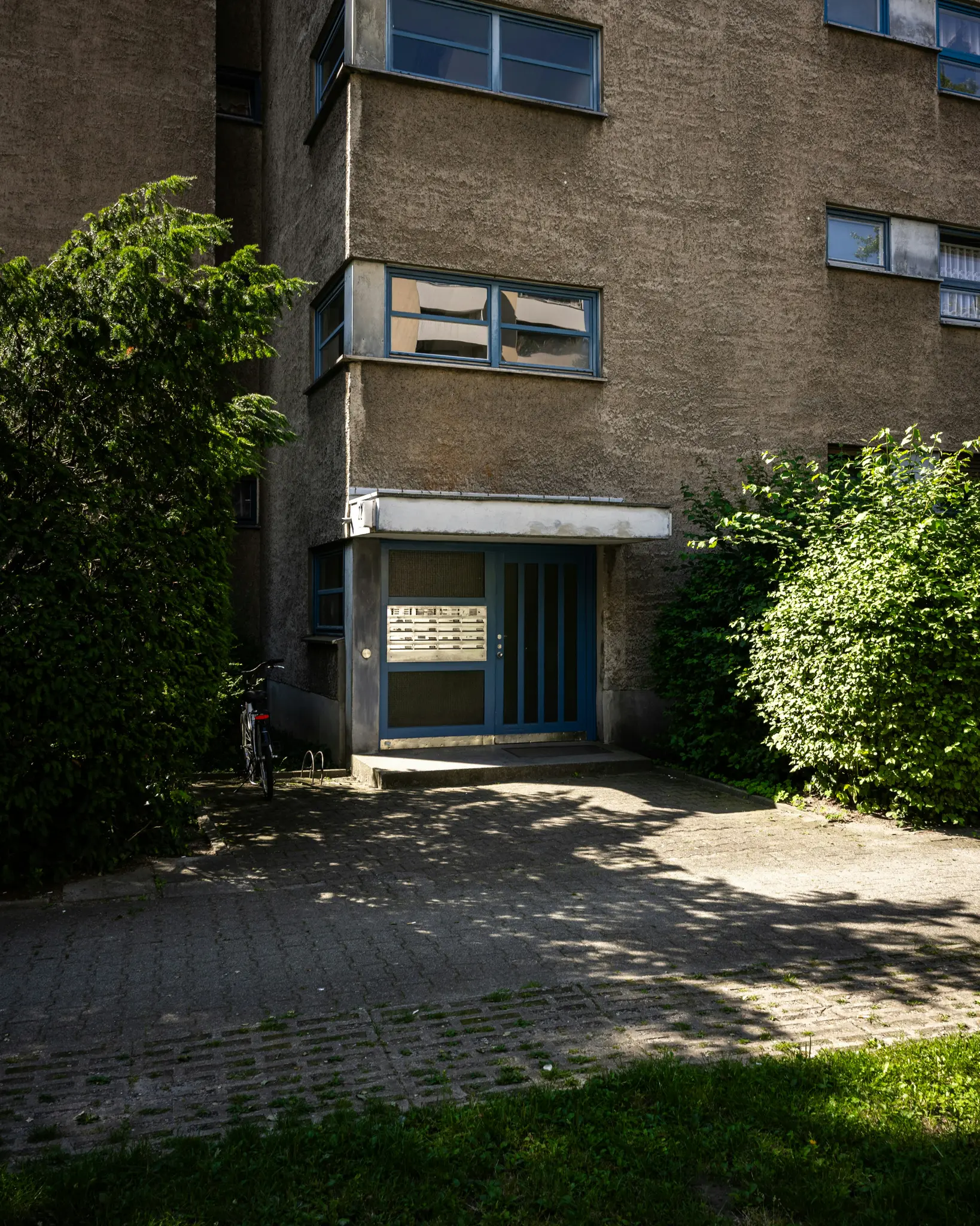Un edificio de hormigón texturizado con ventanas de marco azul y puerta de entrada azul. Arbustos verdes flanquean la