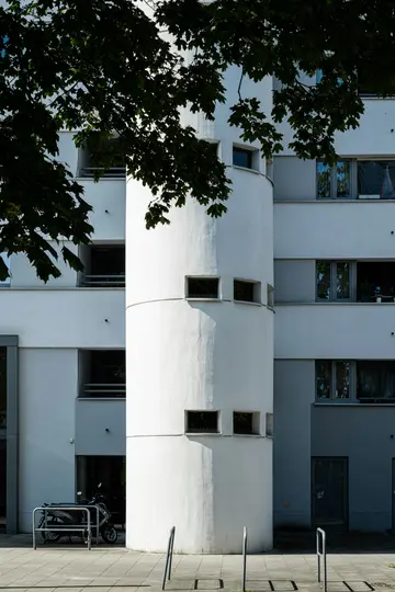 Un moderno edificio bianco e grigio con un'alta sezione cilindrica con piccole finestre rettangolari. Albero verde scuro…
