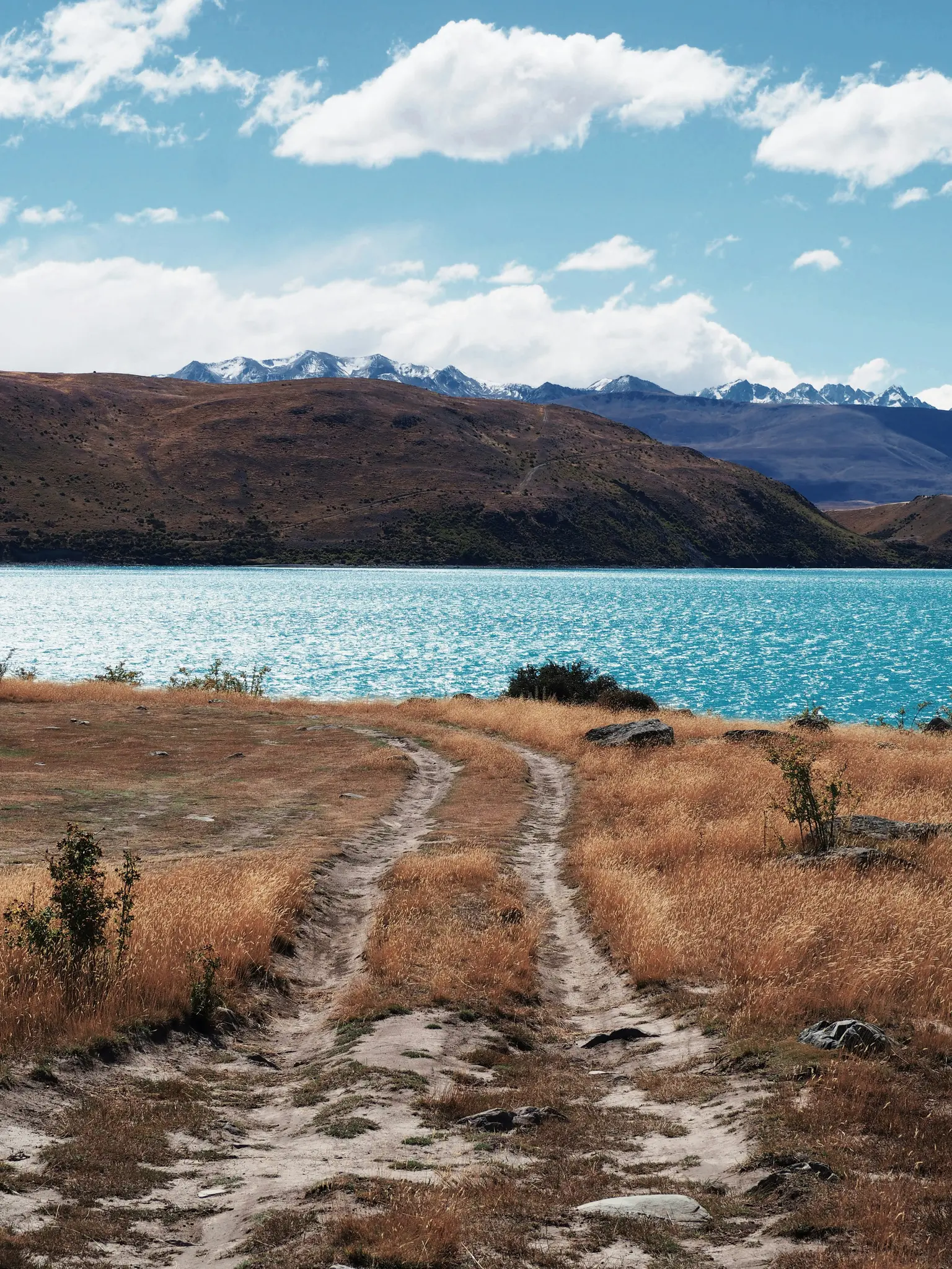 Una pista de tierra serpentea a través de hierba seca y dorada en primer plano, conduciendo hacia un lago de color azul