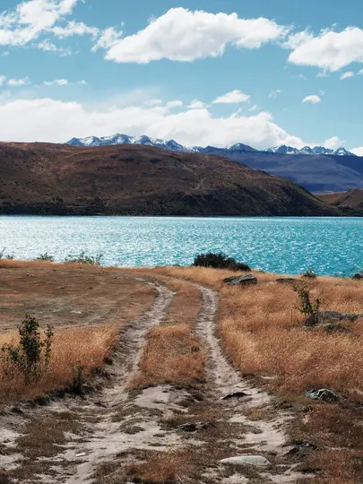 Una pista de tierra serpentea a través de hierba seca y dorada en primer plano, conduciendo hacia un lago de color azul