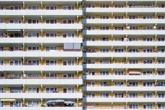 Πλήρης, μετωπική όψη της πρόσοψης μιας πολυώροφης πολυκατοικίας, με σειρές από λευκά μπαλκόνια με κόκκινα κιγκλιδώματα.
