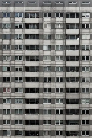 Eine Ganzkörperaufnahme, von vorne, einer hohen, grauen Betonfassade eines Wohngebäudes mit vielen rechteckigen Fenstern und
