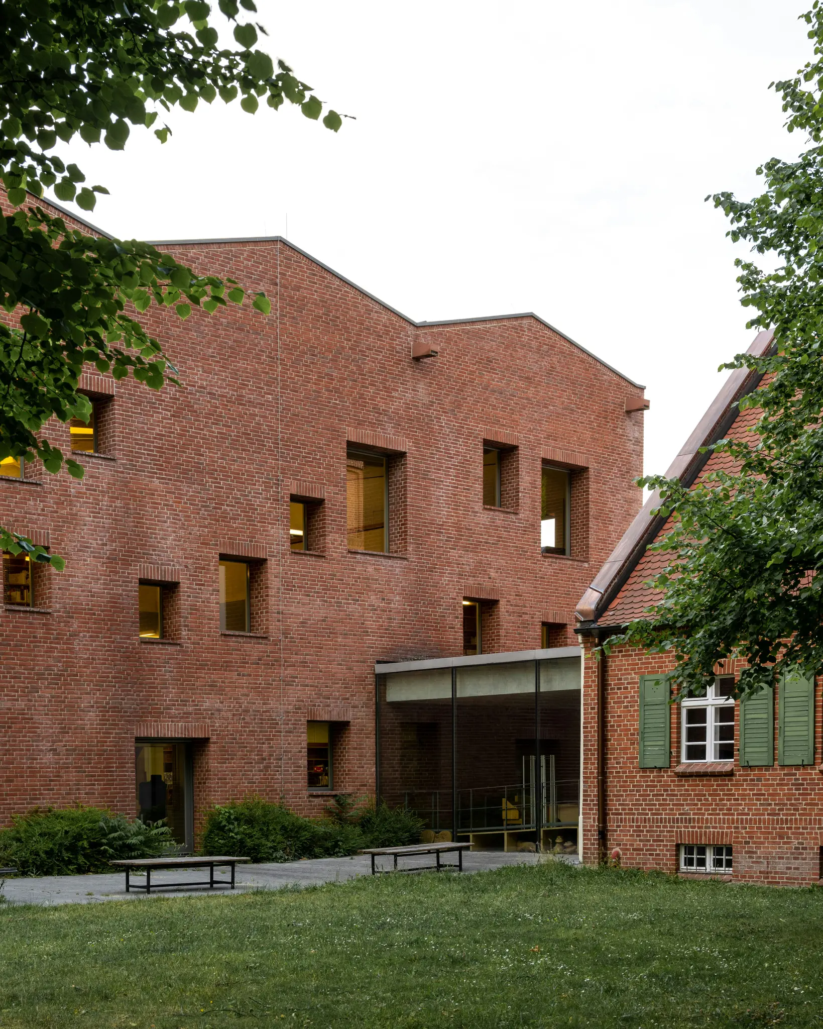 A modern red brick building with a glass entrance connects to an older brick building with a red tiled roof and green…