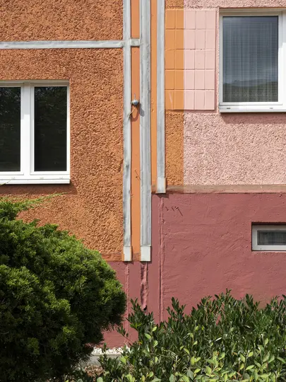Detail exteriéru budovy s částmi oranžové omítky, růžovou texturovanou stěnou a červenohnědým soklem.