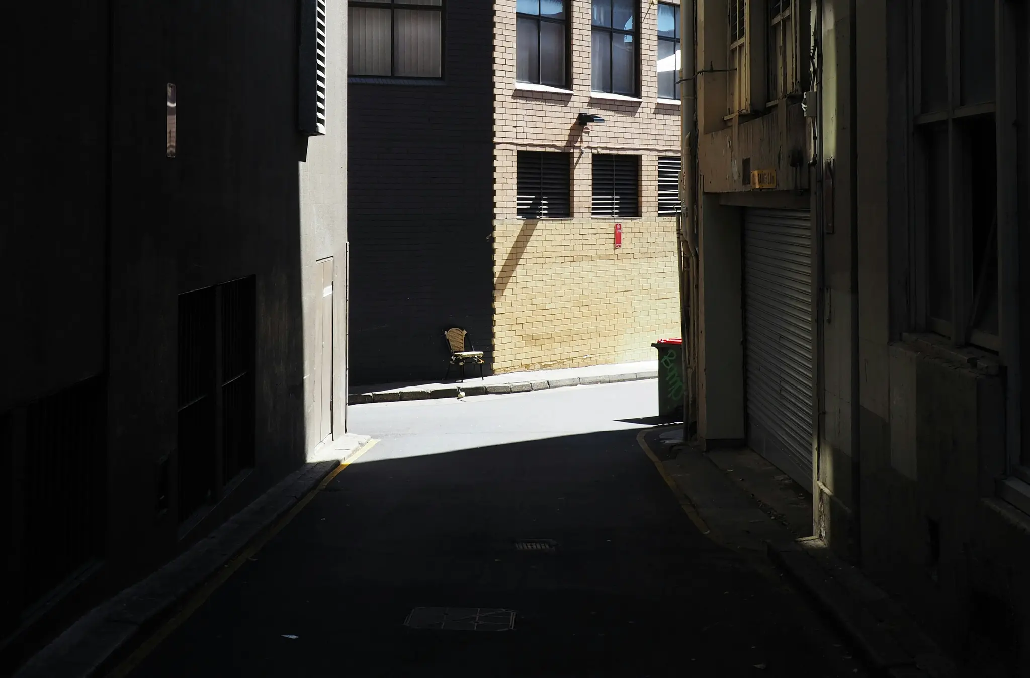 Un callejón estrecho y oscuro con edificios altos a ambos lados. Una única silla de color claro apoyada contra una pared de