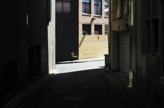 Un vicolo stretto e buio con alti edifici su entrambi i lati. Un'unica sedia chiara appoggiata a un muro di mattoni