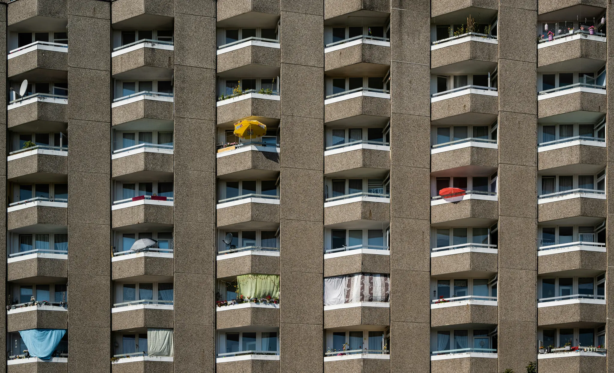Detailný záber fasády betónového bytového domu cez celú snímku. Budova má mnoho balkónov s bielymi zábradliami.