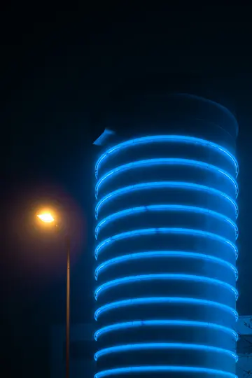 Un edificio alto e cilindrico con luci al neon orizzontali blu brillante avvolte all'esterno, accanto a un...