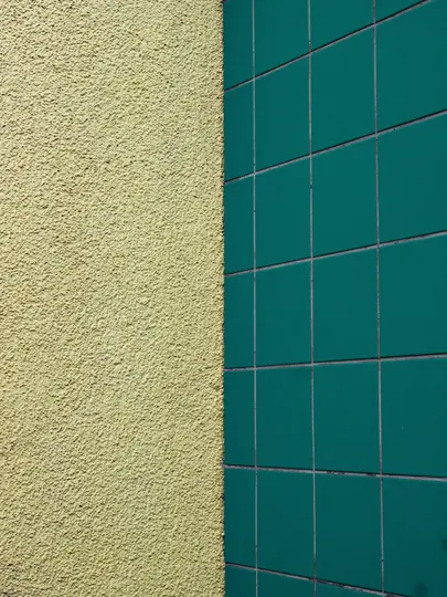 Lähikuva, abstrakti näkymä rakennuksen kulmasta, vasemmalla keltainen rappausseinä ja oikealla vihreillä laatoilla