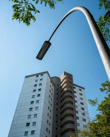 Záber z nízkeho uhla na vysoký, sivý obytný dom s výraznou zakrivenou časťou balkónov. Pouličná lampa sa zakrivuje…