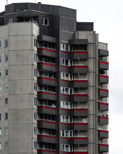 Vysoký, brutalistický bytový dům se šedými betonovými zdmi a tmavě šedými balkony s červenými pruhy.