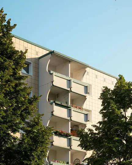 Un edificio color crema con più balconi, parzialmente nascosto da lussureggianti alberi verdi su entrambi i lati, sotto un