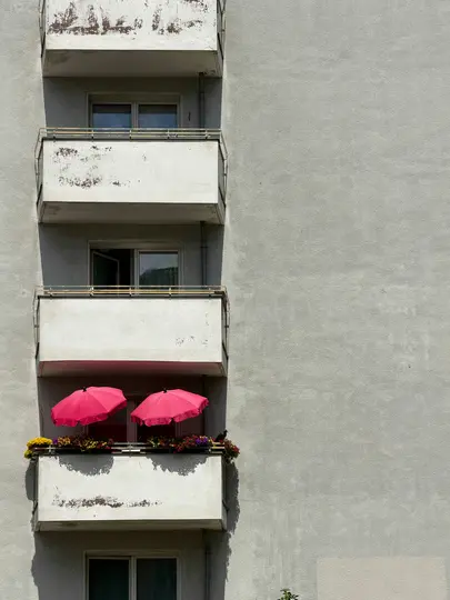 Un primo piano della facciata in cemento grigio di un condominio con diversi balconi. Un balcone presenta due ombrelli rosa