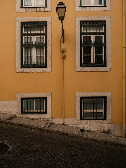 Facciata di un edificio giallo con quattro finestre, due con grate verde scuro decorate e un lampione nero. Una strada