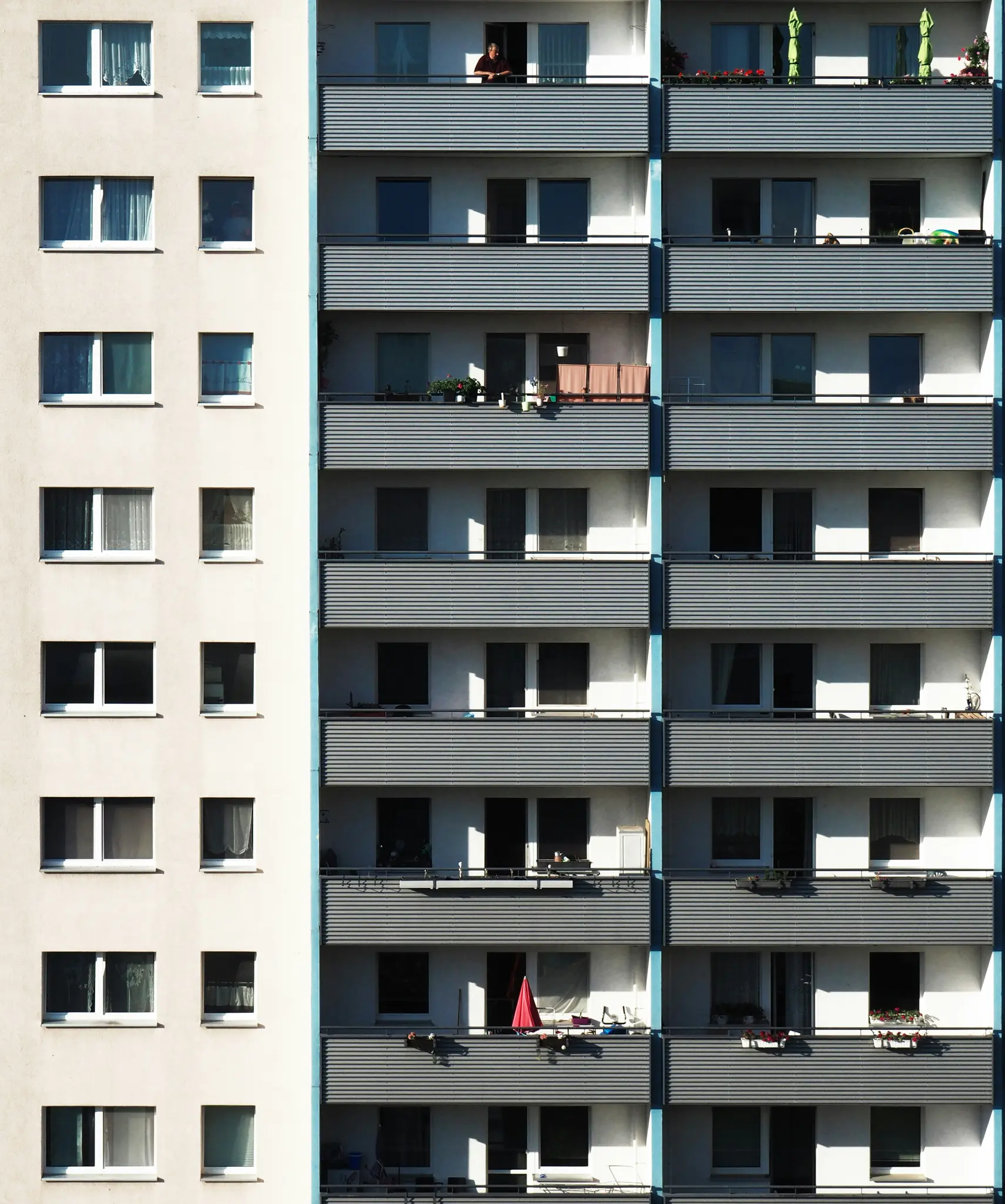 Una alta fachada de edificio de apartamentos con hileras de ventanas y balcones. Una persona se apoya en la barandilla de un