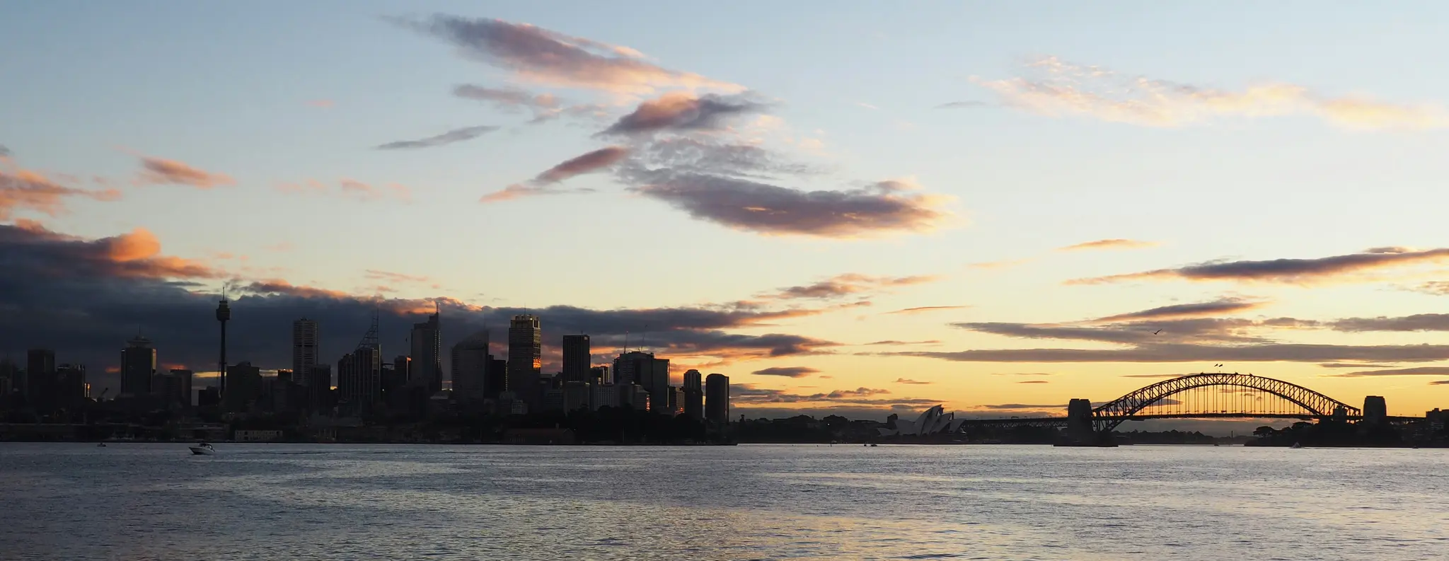 Una foto panorámica del skyline de Sídney y el Puente Harbour perfilados contra un cielo al atardecer con tonos naranjas y