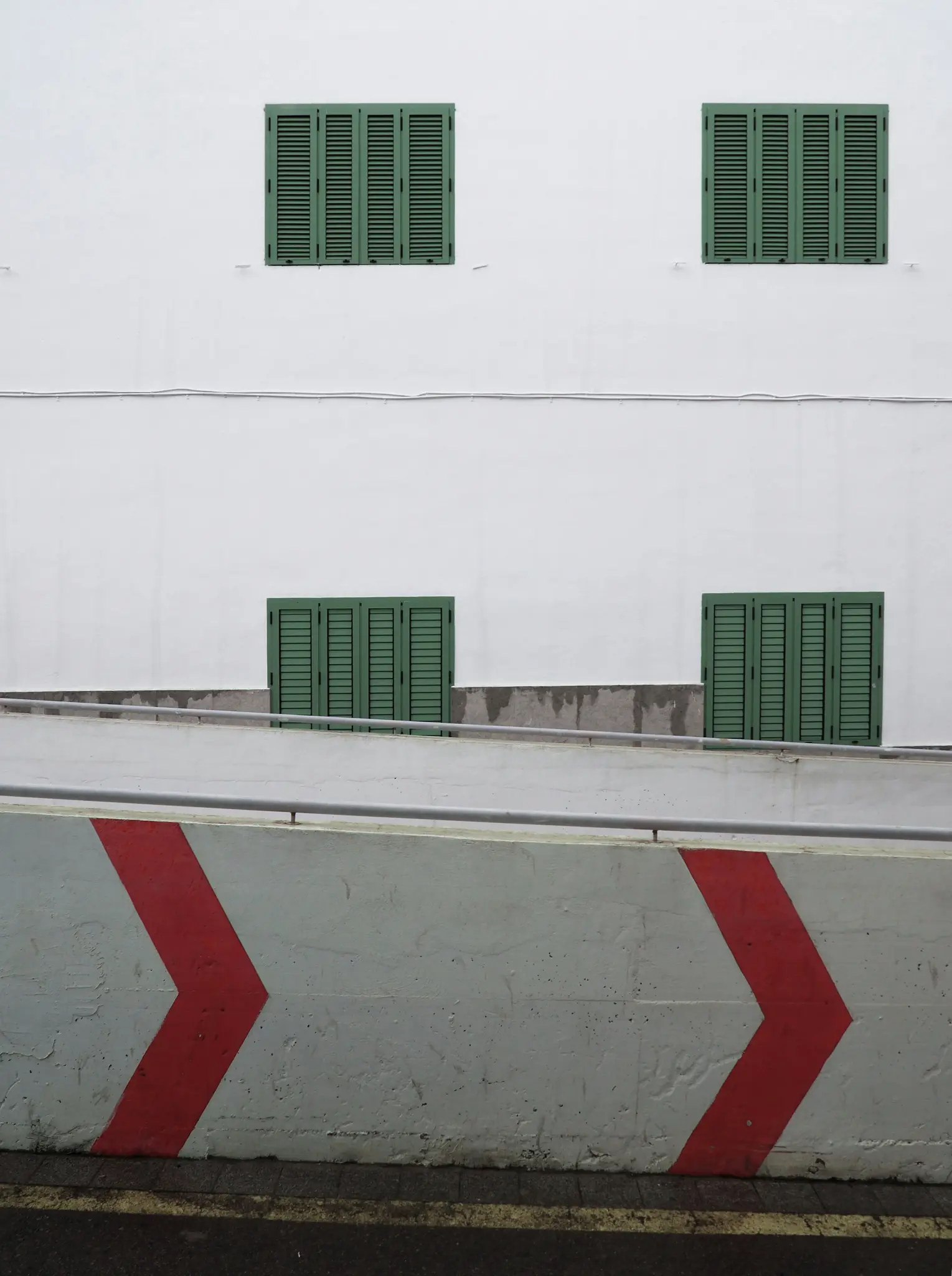 Una fachada blanca de edificio tiene cuatro contraventanas de color verde oscuro. Debajo, una rampa de hormigón tiene un