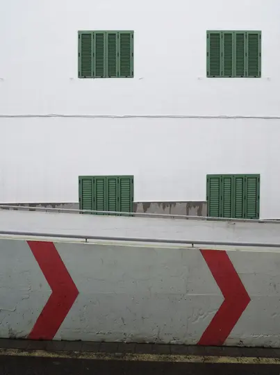 Eine weiße Gebäudefassade mit vier dunkelgrünen Fensterläden. Darunter hat eine Betonrampe ein rotes Chevronmuster, das nach…