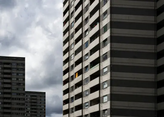Tre edifici brutalisti con molte finestre e balconi sotto un cielo grigio e nuvoloso. Gli edifici sono grigi e…