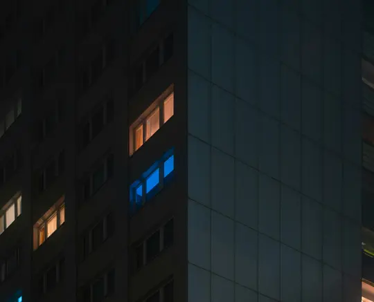 Una fachada oscura de edificio por la noche con varias ventanas que brillan con cálida luz naranja y una ventana que brilla