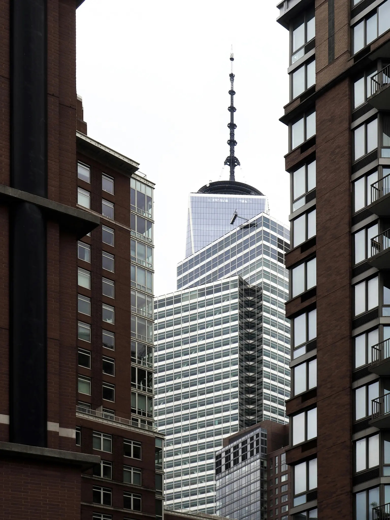 Una aguja alta y oscura del One World Trade Center se eleva contra un cielo blanco, enmarcada por edificios de ladrillo