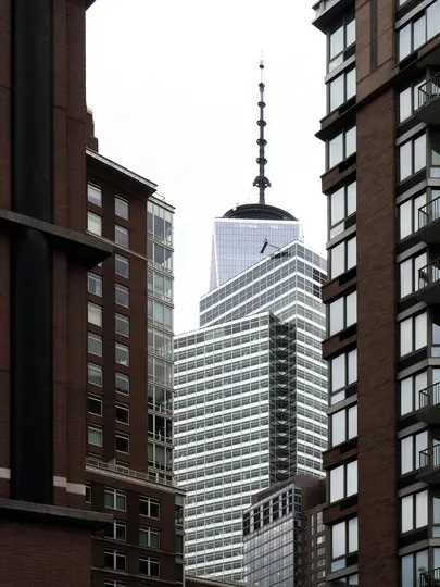 Un'alta e scura guglia del One World Trade Center si erge contro un cielo bianco, incorniciata da edifici in mattoni marroni