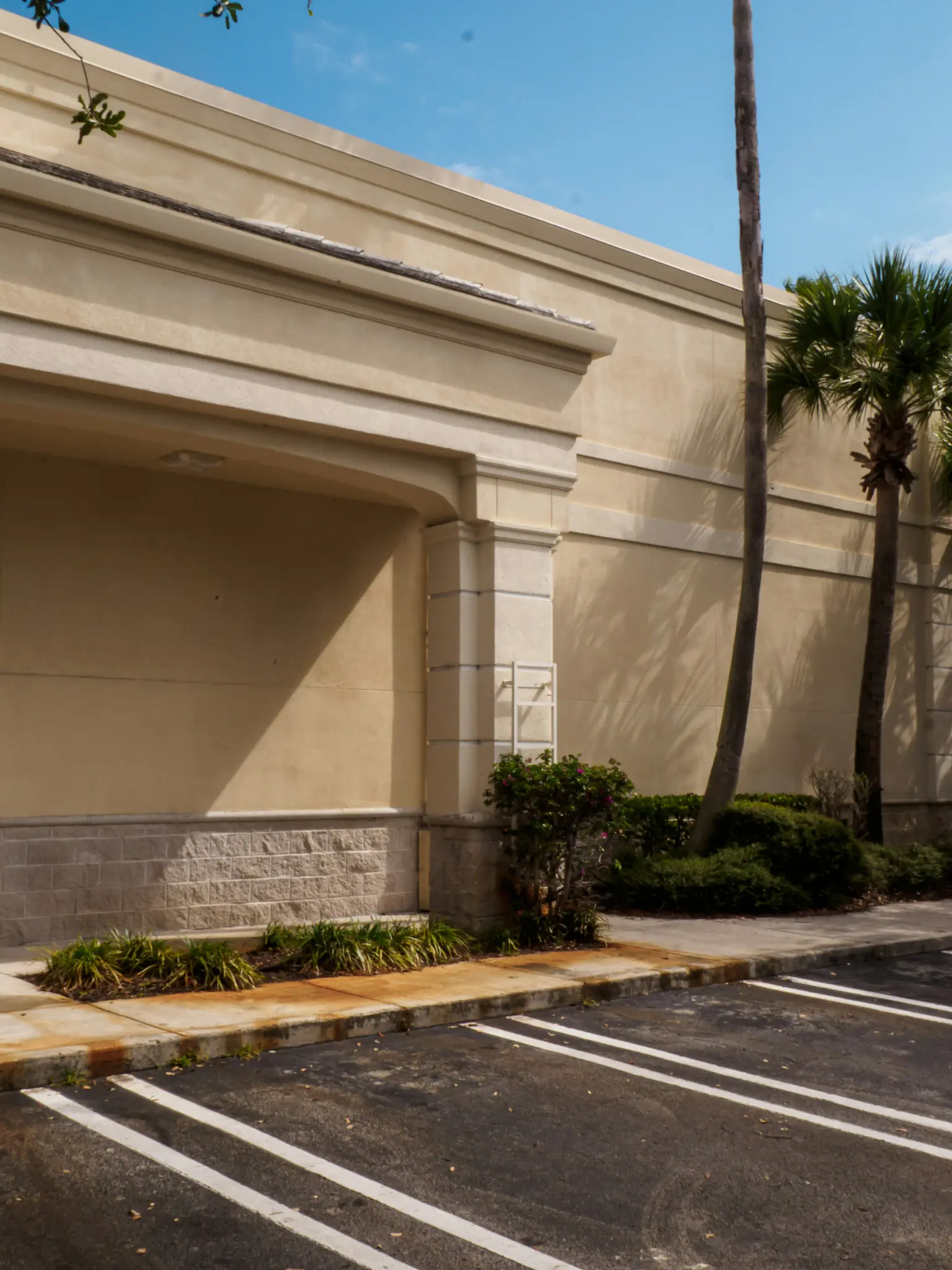 Un edificio comercial beige con una base de piedra gris y una entrada arqueada. Un aparcamiento de asfalto negro con líneas