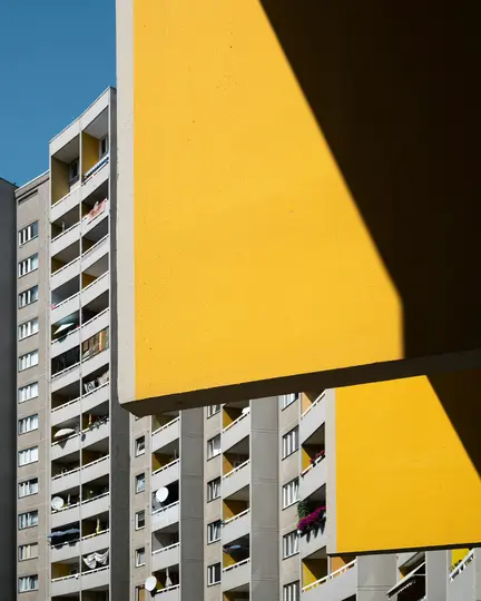 Jasne žltá fasáda budovy s ostrými tieňmi v popredí, čiastočne zakrývajúca vysoký sivý bytový dom.