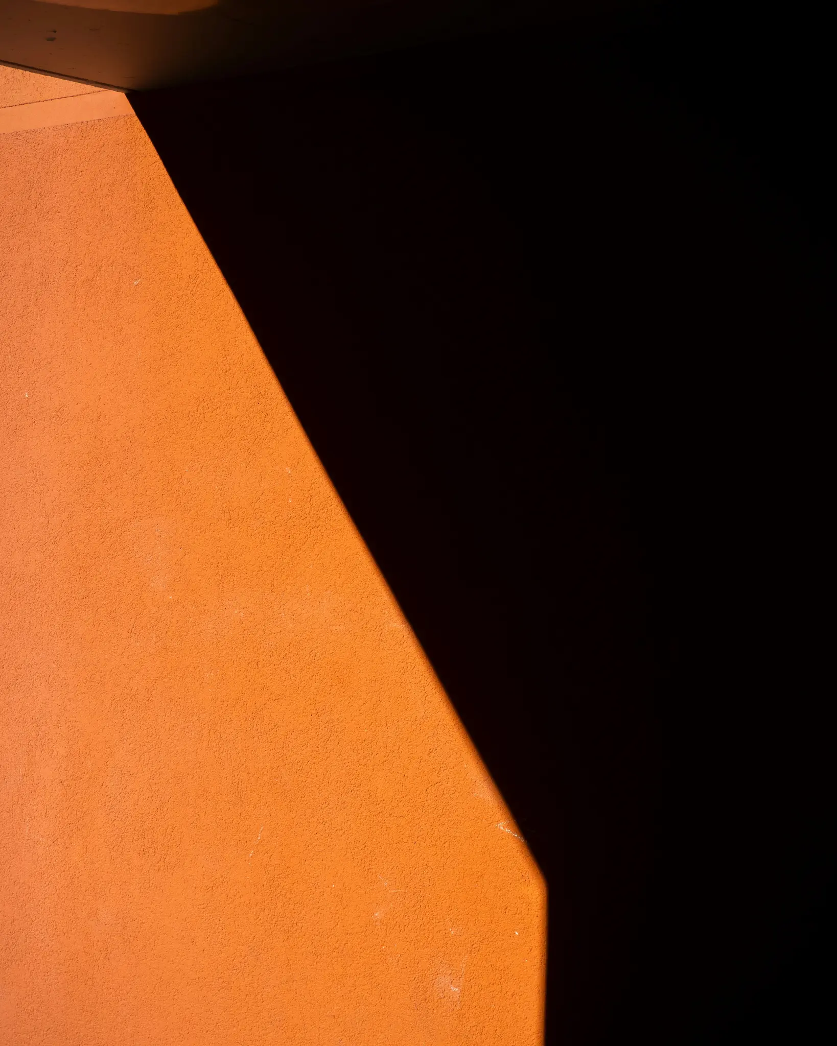 Striedma, abstraktná fotografia oranžovej textúrovanej steny ostro rozdelenej diagonálnym tieňom, vytvárajúca silný kontrast.