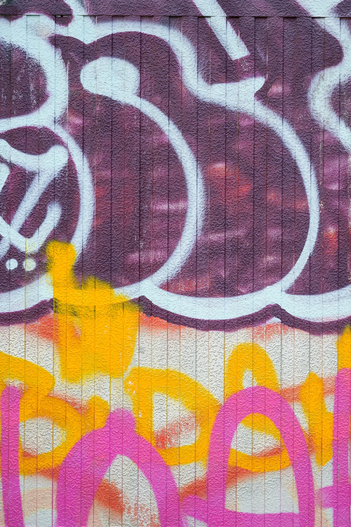 Detailný záber na textúrovanú stenu pokrytú viacerými vrstvami graffiti vo fialovej, bielej, žltej a ružovej farbe.