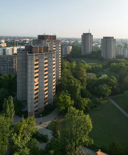 Letecká fotografia zobrazuje niekoľko vysokých, sivých obytných budov s balkónmi, pretkaných sviežimi, zelenými stromami a...