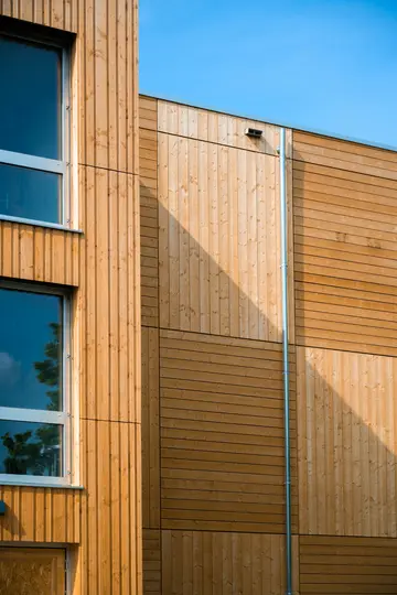 Un primo piano della facciata di un edificio moderno con pannelli in legno marrone chiaro e scuro, due finestre e un pluviale