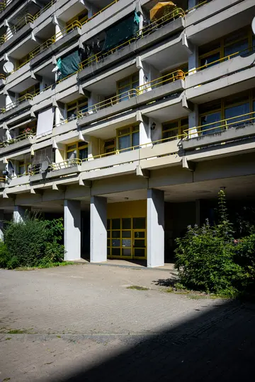 Vícepodlažní betonový bytový dům s mnoha balkony se žlutými zábradlími a rámy oken. Přízemí...