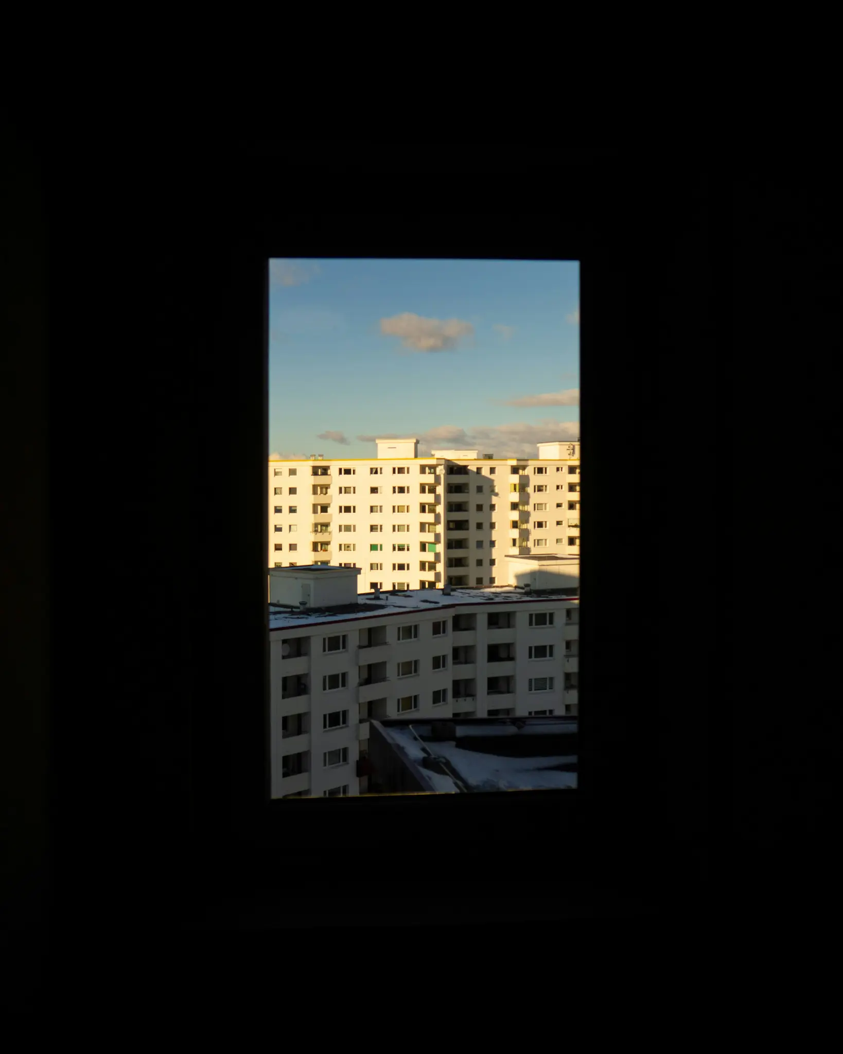 Una vista rectangular a través de una ventana oscura muestra varios edificios de apartamentos blancos bajo un cielo azul con